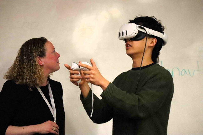 A woman and a young man stand in front of a whiteboard. The man is wearing a VR headset and holding two VR controllers in his hands.