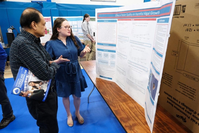 A young woman and a man stand in front of a poster set on a table.