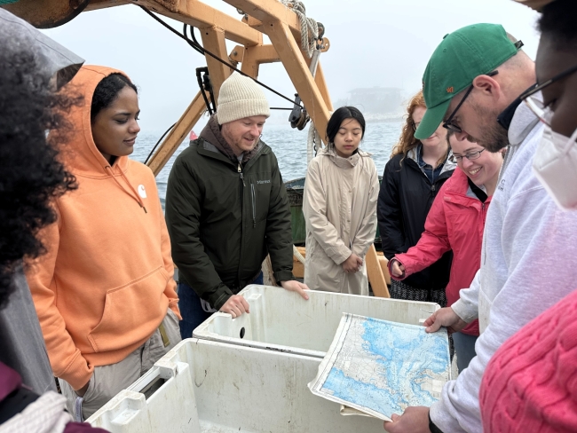 People gather in a circle around empty totes on a boat and look at a map.
