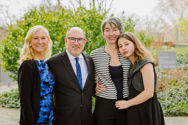 Three women and a man stand in front of some shrubbery.