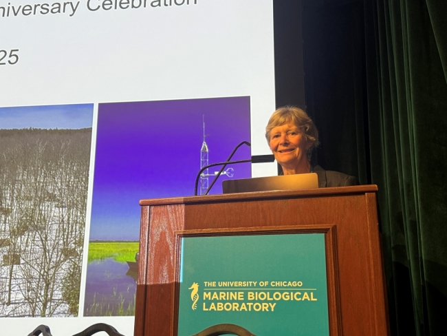 A woman stands behind a podium with a microphone and smiles. A large screen is partially visible behind her.