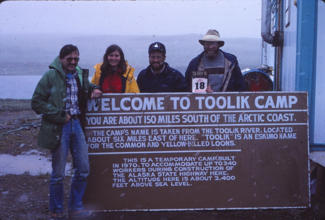 Four people pose behind/in front of a sign that says "Welcome to Toolik Camp". A wide expanse of land and water is visible behind them, and a building is next to them.