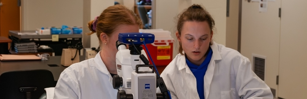 Two scientists in white coats are seated behind a microscope.