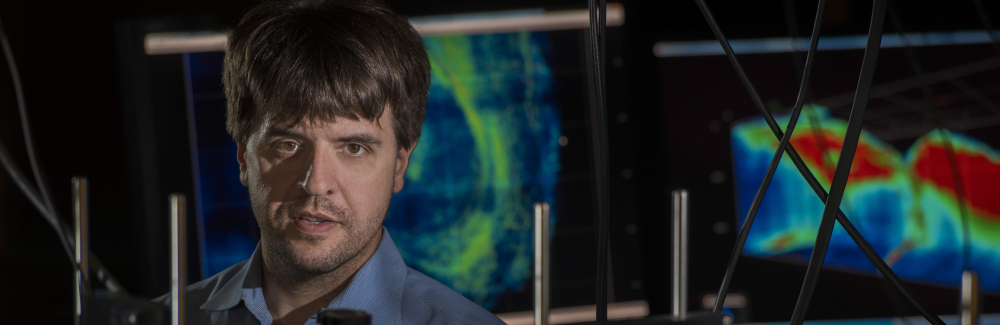 Karl Desseroth in lab behind microscopes with flourscent microscopy images in background 