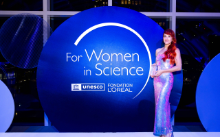 Kaitlyn Webster wearing a gown, holding an award, standing to the right of a "For Women in Science" sign. 