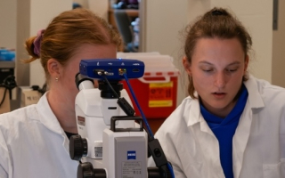 Two scientists in white coats are seated behind a microscope.
