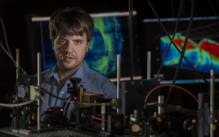 Karl Desseroth in lab behind microscopes with flourscent microscopy images in background 