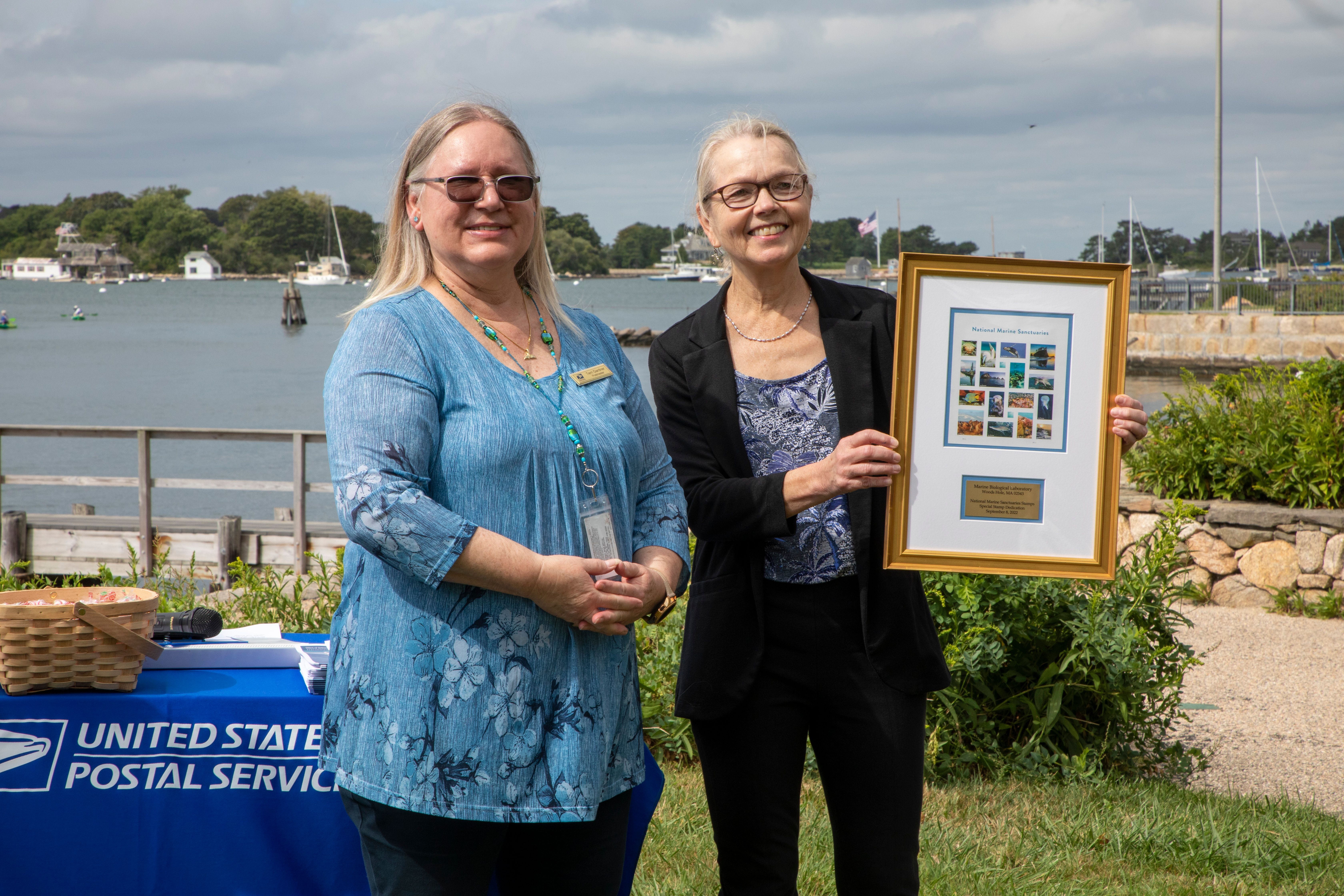Sylvester stands with Theresa Gardner, West Falmouth Postmaster, and holds a print of the National Marine Sanctuary forever stamps. Credit: © Woods Hole Oceanographic Institution