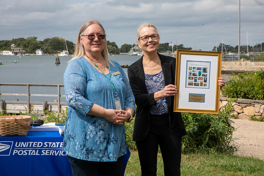 Sylvester stands with Theresa Gardner, West Falmouth Postmaster, and holds a print of the National Marine Sanctuary forever stamps. Credit: © Woods Hole Oceanographic Institution