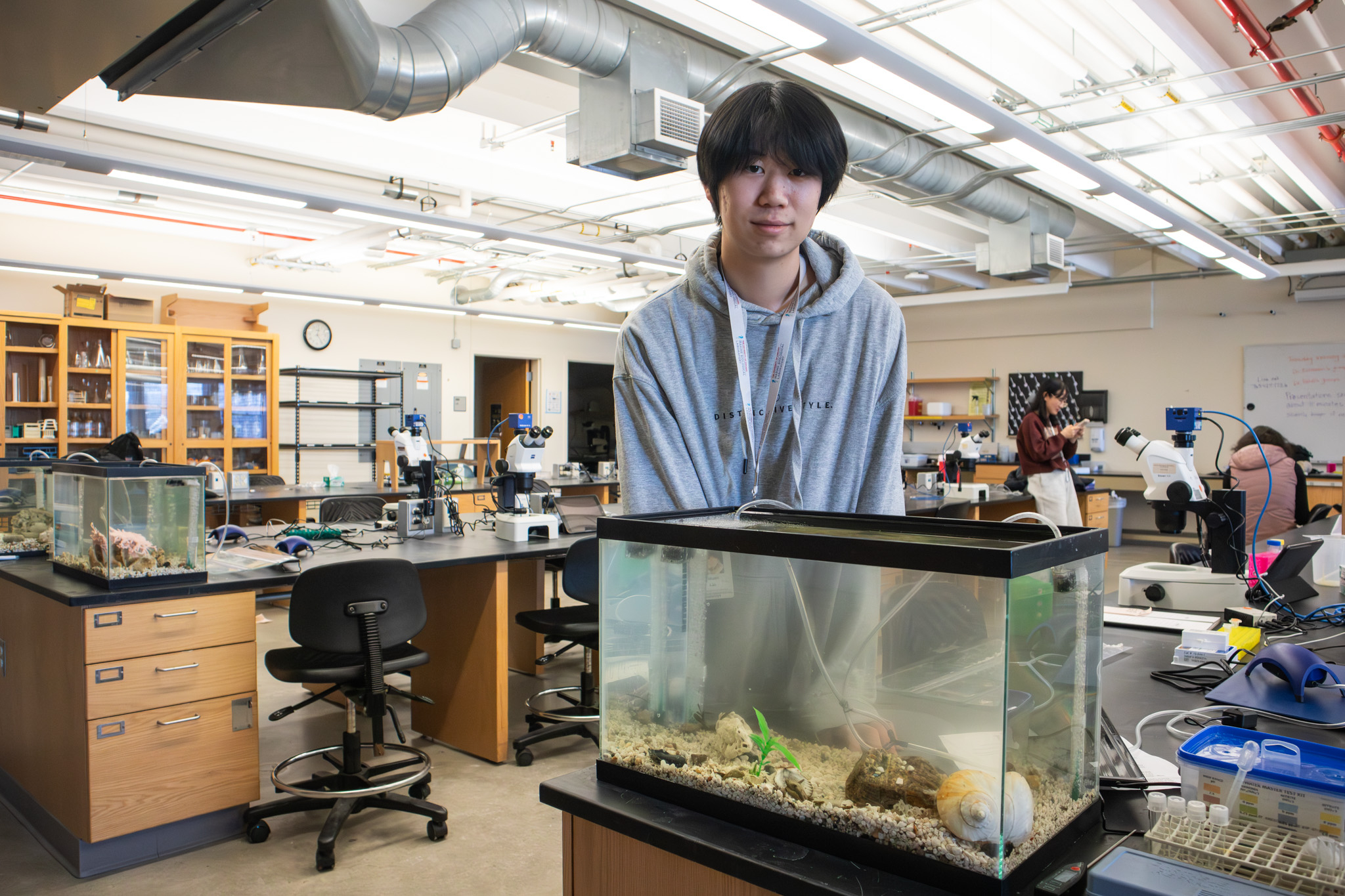 This photo shows a high school student looking into the camera and standing behind a fish tank in a science lab.
