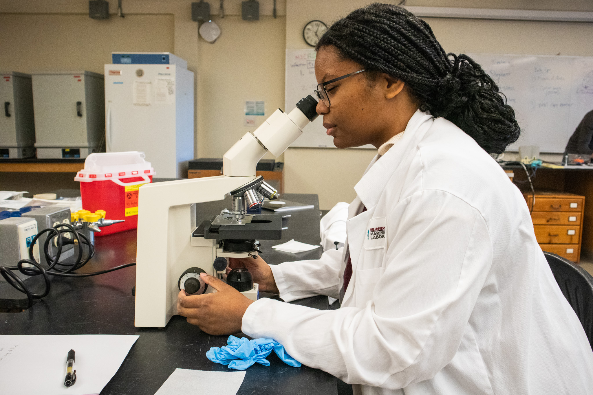This photo shows a high school student wearing a white lab coat peering into a microscope in a science lab.