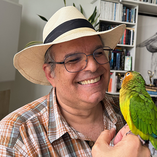 Smiling man wearint a light straw hat and plaid shirt, holding a green and yellow parrot.