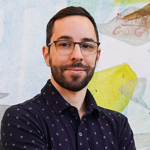 Man with dark hair and beard wearing glasses and a dark patterned shirt.