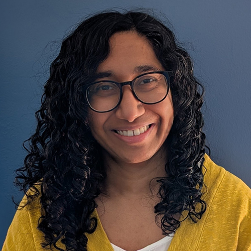 Smiling woman with long, curly dark hair wearing glasses and a white shirt with yellow jacket.
