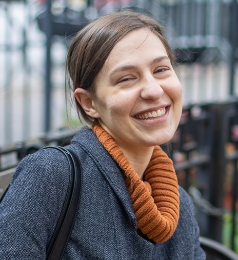Smilling woman with dark hair wearing a dark orange sweater and gray jacket.