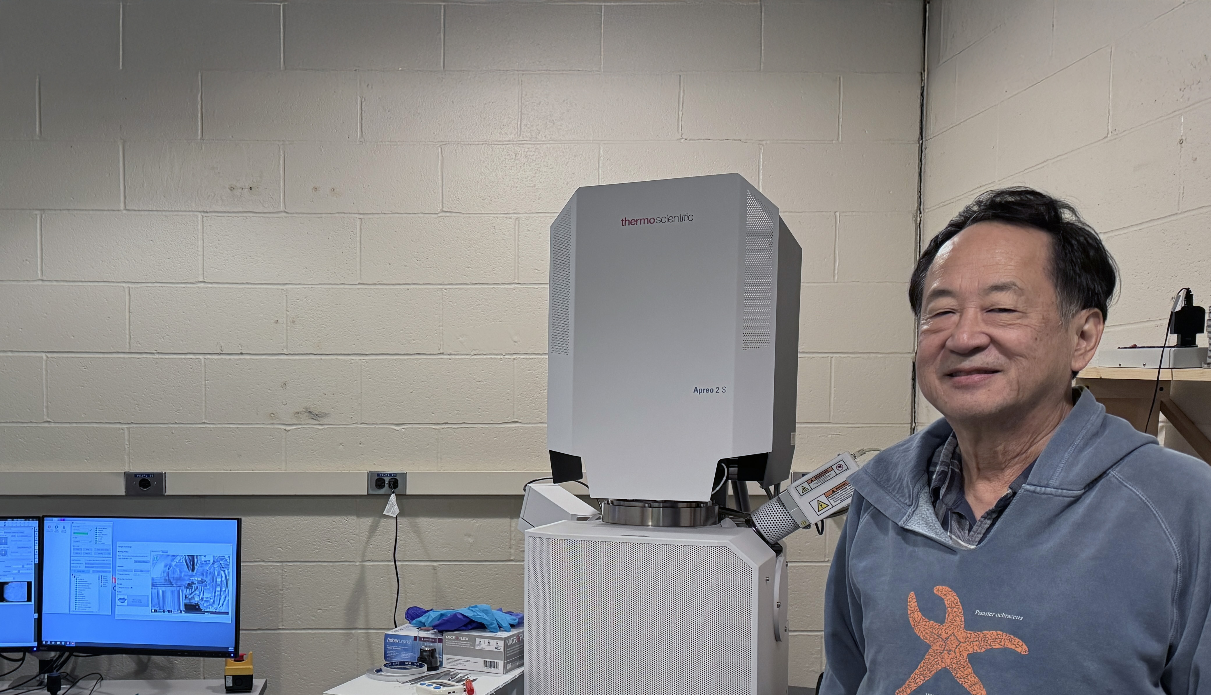 Smiling man with dark hair and blue hoodie in a lab with a computer and equipment.