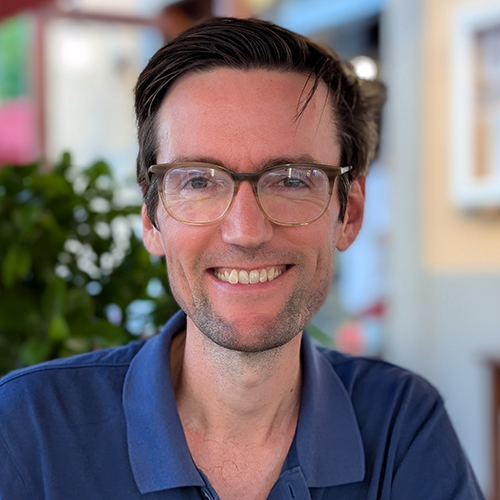 Smiling man with short dark hair, wearing glasses and a blue collared shirt.