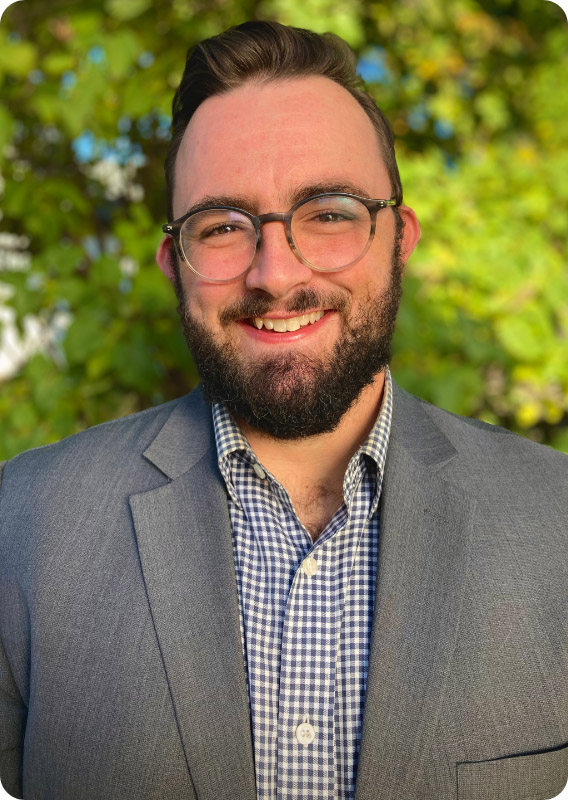 Smiling man with dark hair and beard, wearing glasses and checkered shirt with gray coat. He is standing in front of a bright green tree.