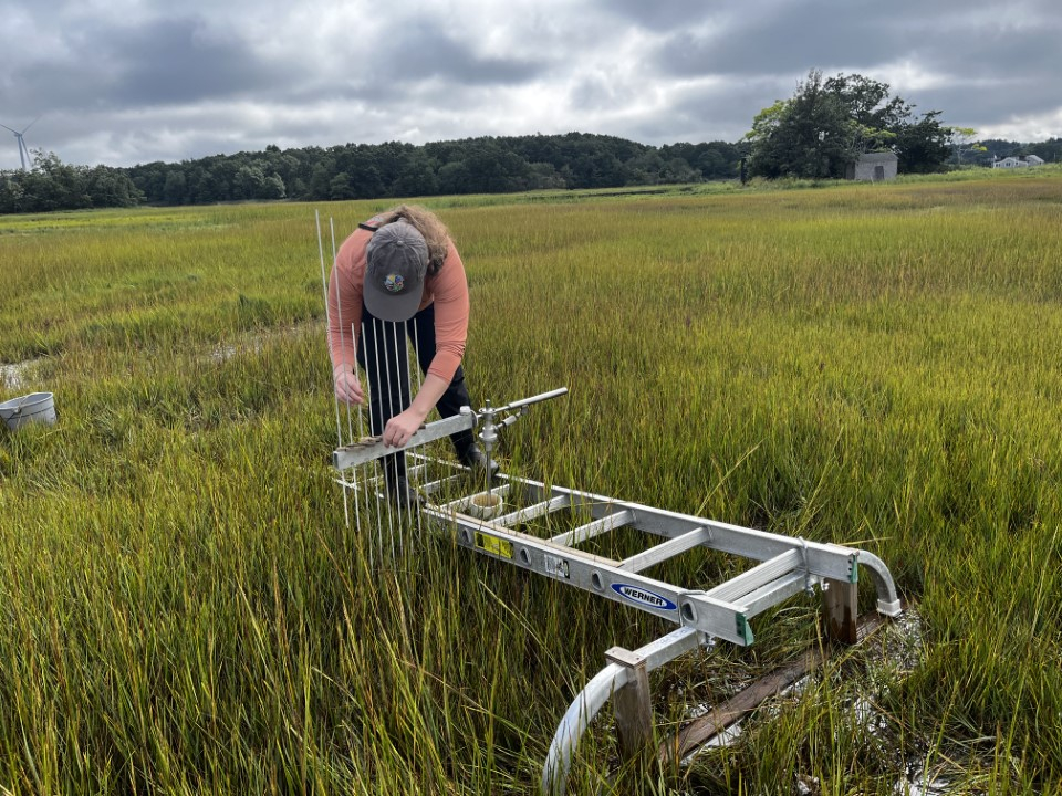 This photo shows a woman in a field bending down to do work on a metal contraption.
