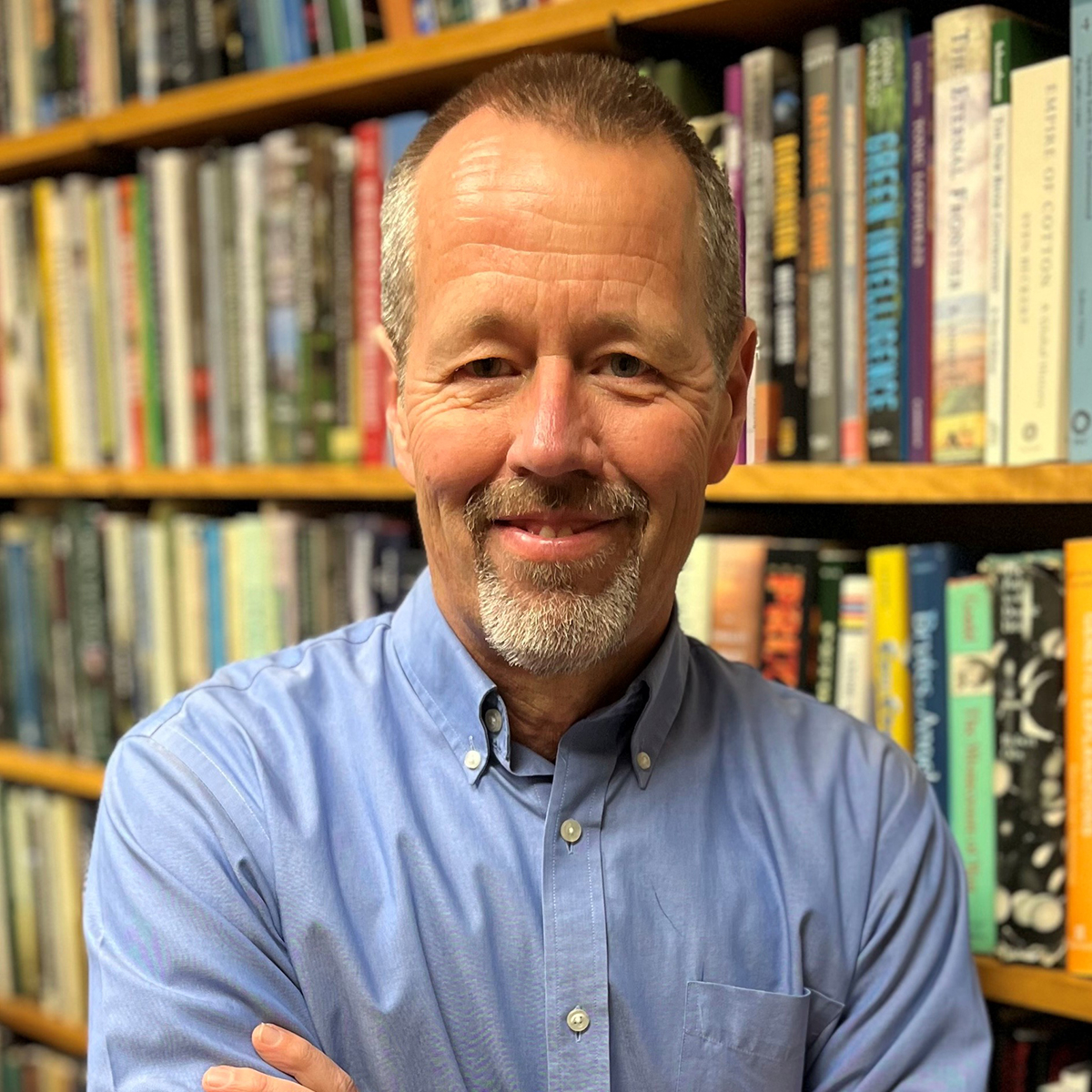 Smiling white man with short graying hair and beard, wearing a blue shirt and standing in front of a bookshelf.