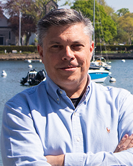 Male presenting person with short hair, standing in front of the water with boats in the background,