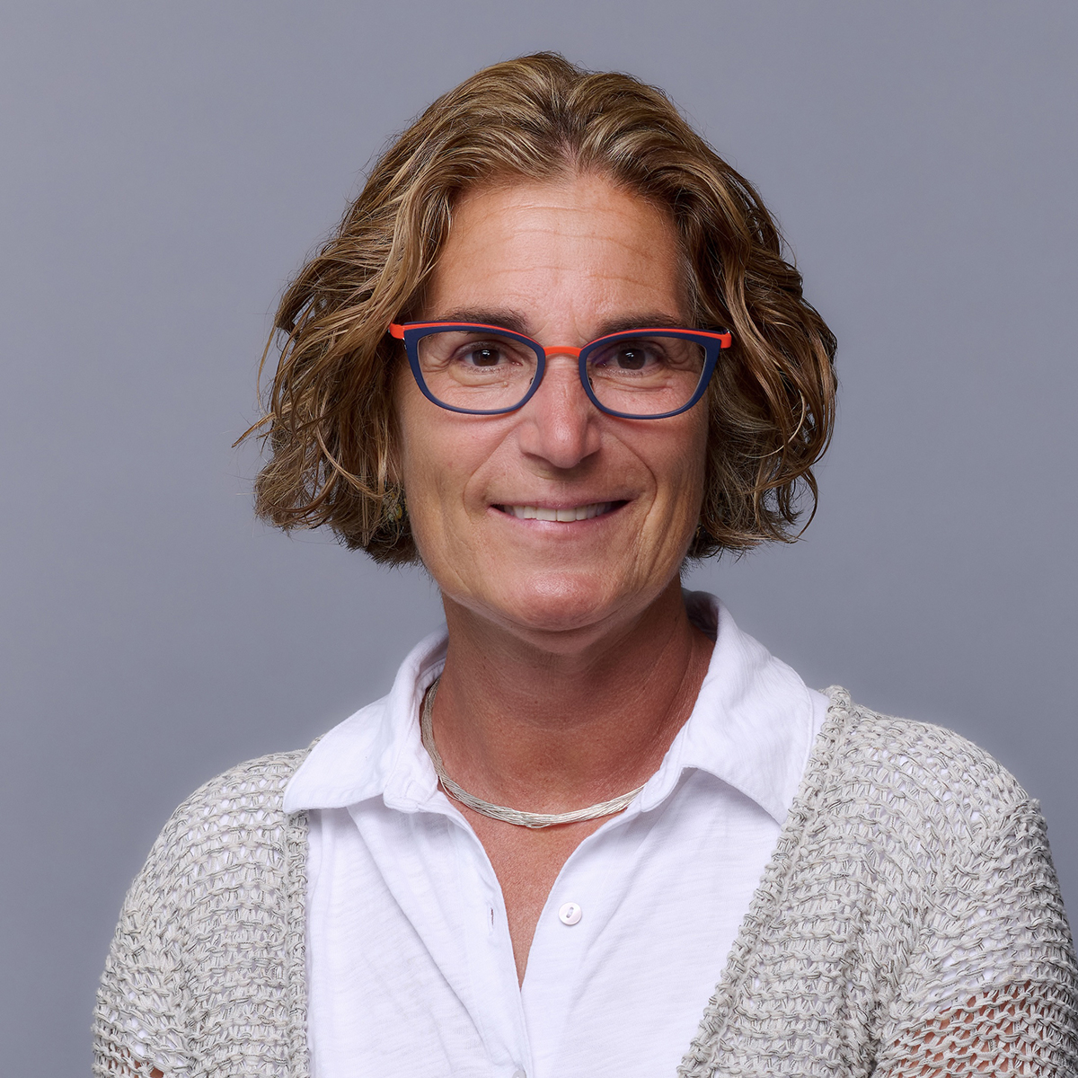 Smiling white woman with short light blond hair, wearing red glasses and a white shirt with brown sweater.