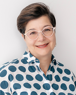 Female presenting person with short dark hair and glasses, wearing a blue and white polka dot shirt.