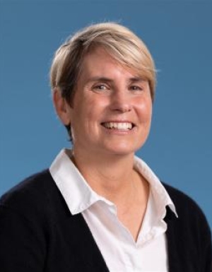 Jen Walton smiles at the camera. She wears a white collared shirt with a black cardigan. Her hair is short and blonde. She sits in front of a blue background.
