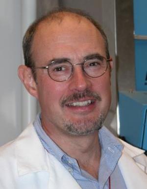 Karl Matlin smiles at the camera. He wears glasses, a blue collared shirt, and a white lab coat.