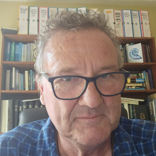 Male presenting person with short gray hair, wearing glasses and sitting in front of a bookshelf.