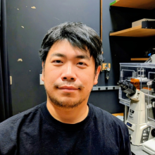 Male presenting person with short dark hair standing in front of a microscope.