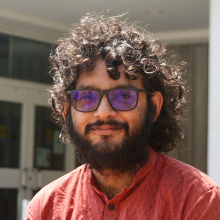 Male presenting person with dark curly hair and beard wearing sunglasses.