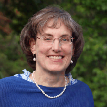Female presenting person with short brown hair wearing glasses, a blue sweater and pearls.
